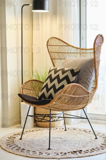 A cozy hotel room features a stylish wicker chair adorned with chevron-patterned and plain cushions. Soft lighting and natural textures create a relaxing bohemian ambiance
