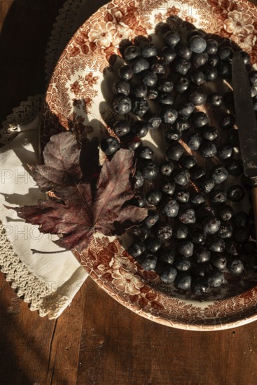 A rustic autumn still life featuring fresh blueberries on a vintage decorative plate. A dry maple leaf and a lace doily add a cozy, nostalgic touch to the wooden table setting