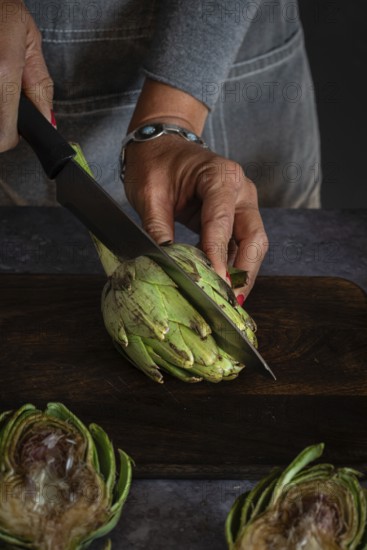 A person in a gray apron skillfully cuts an artichoke with a large kitchen knife on a dark wooden board. The scene captures the essence of fresh, healthy cooking