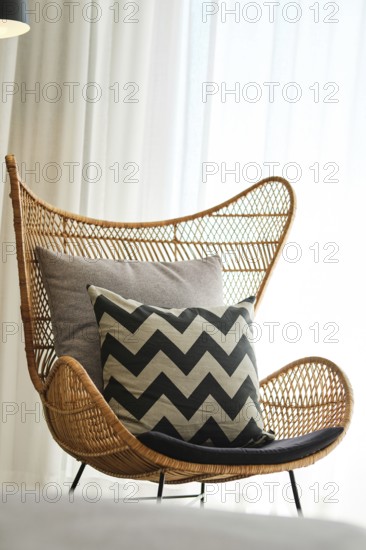 A stylish rattan chair adorned with cushy grey and black chevron pillows sits beside sheer curtains, creating a cozy and modern hotel room ambiance
