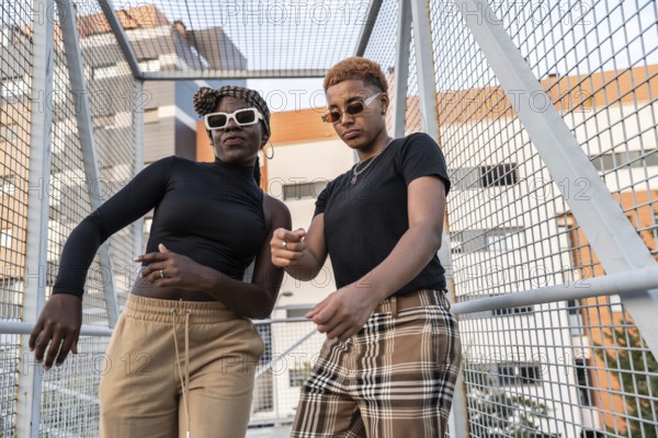 A vibrant LGBT multiethnic couple poses joyfully in a modern urban setting, exuding confidence and style against a backdrop of city buildings and metal structures