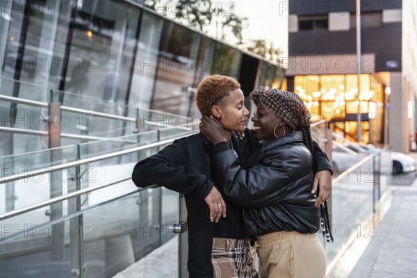 An affectionate LGBT multiethnic couple embraces outdoors in the city, capturing a candid and heartfelt moment. They are surrounded by modern buildings and soft evening lighting