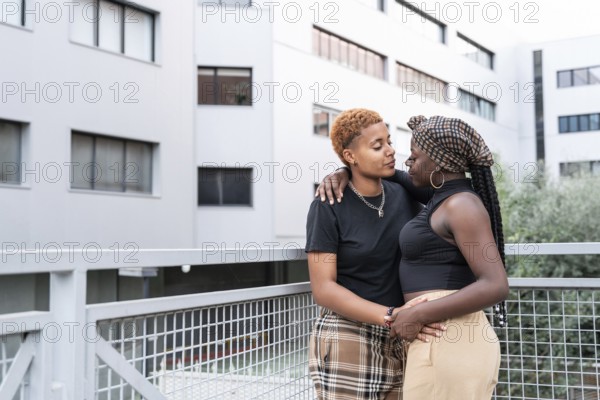 An affectionate LGBT multiethnic couple leans against a balcony railing in an urban setting, sharing a loving gaze. The backdrop is a modern building, adding urban sophistication to the moment