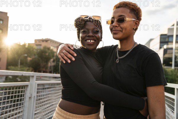 An LGBT multiethnic couple embraces warmly on a pedestrian bridge. They smile with genuine happiness, surrounded by city buildings. The sun sets in the background, casting a warm glow