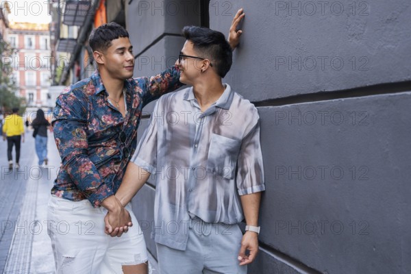 A smiling latin gay couple holds hands and enjoys a sunny day in the streets of Madrid, surrounded by historic buildings and a vibrant urban atmosphere