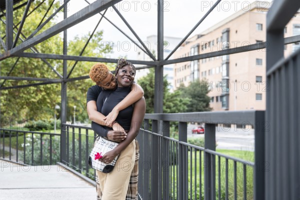 An affectionate LGBT multiethnic couple embraces on a city bridge, radiating happiness and love. Urban buildings and greenery in the background create a warm, modern atmosphere