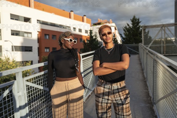 A stylish LGBT multiethnic couple poses confidently on an urban bridge under the warm, golden sunlight. Both wear sunglasses and casual attire, expressing joy and togetherness