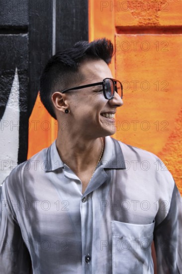 A stylish latin man wearing sunglasses and a patterned shirt smiles broadly against a vibrant orange wall background, exuding joy and confidence