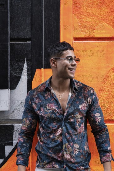 A stylish latin man in a floral shirt and sunglasses stands smiling against a vibrant orange and black graffiti wall, embodying urban fashion and contemporary style