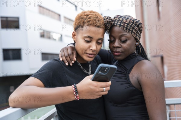 A loving multiethnic couple stands close, sharing a moment while looking at a smartphone. They embrace and smile, enjoying each other's company in a vibrant urban environment