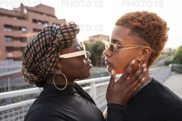 An LGBT multiethnic couple gazes into each other's eyes on a sunlit urban bridge, showcasing intimacy and style. Their expressions capture a moment of genuine affection and connection