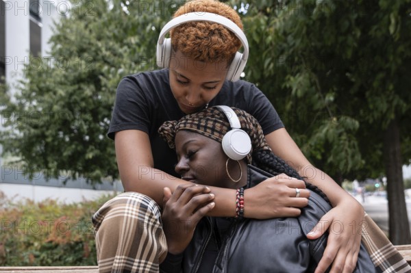 An affectionate multiethnic LGBT couple, embracing outdoors, share a tender moment while both wearing headphones. They enjoy the music together, creating a sense of closeness and intimacy