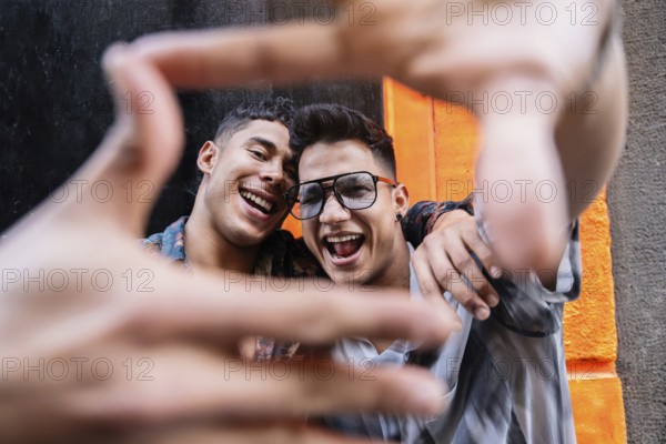 A smiling couple frames their joyful expressions with their hands against a vibrant orange urban backdrop. Their happiness and connection are vividly displayed