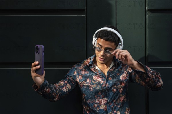 A trendy latin man in floral shirt and sunglasses takes a selfie outdoors, showcasing modern style with headphones on. The photo captures confidence and contemporary fashion