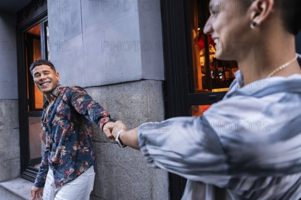 A smiling latin gay couple holds hands outside an urban cafe, radiating happiness and love. One partner, in a floral shirt, winks while being affectionately pulled forward by the other