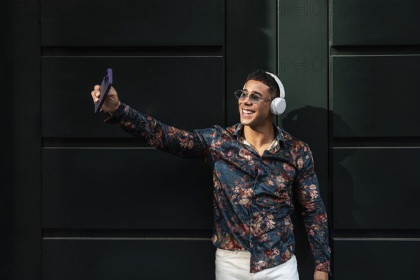 A young latin man in a floral shirt, wearing sunglasses and headphones, smiles while taking a selfie against a dark green wall. The sun highlights his cheerful expression