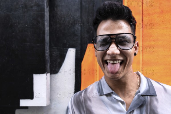 A young latin man wearing oversized sunglasses sticks out his tongue in a playful manner while standing in front of a vibrant orange and black wall, embodying a carefree vibe