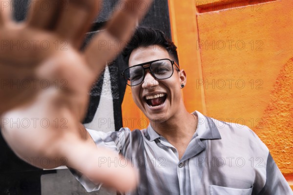 A young man wearing glasses and a stylish shirt smiles joyfully, extending his hand toward the camera. The vibrant orange background enhances the lively, fun atmosphere