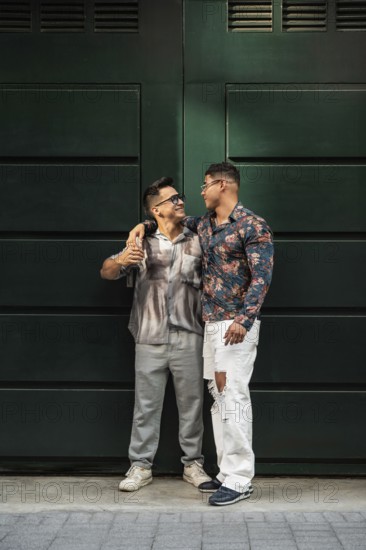 A stylish latin gay couple stands close, exchanging affectionate looks against a large dark green door. Their joyful expressions convey love and connection