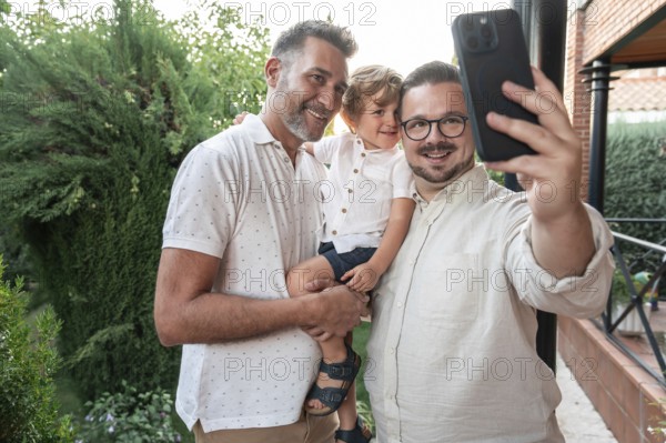 A joyful gay couple takes a smiling selfie with their son in a lush garden setting. The image captures a moment of family love and diverse parenting