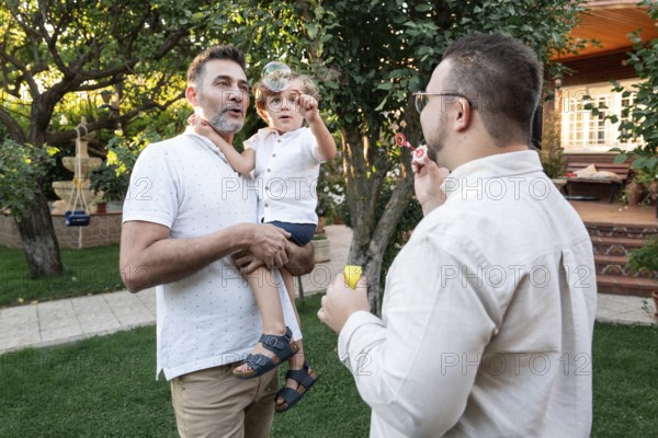 A gay couple enjoys a sunny afternoon in the garden with their young son, creating bubbles and laughter. The family shares a moment of joy under the trees