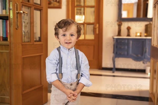 A young boy stands confidently dressed in a light blue shirt with suspenders. He is in an elegantly furnished room with wooden doors and a blue console table