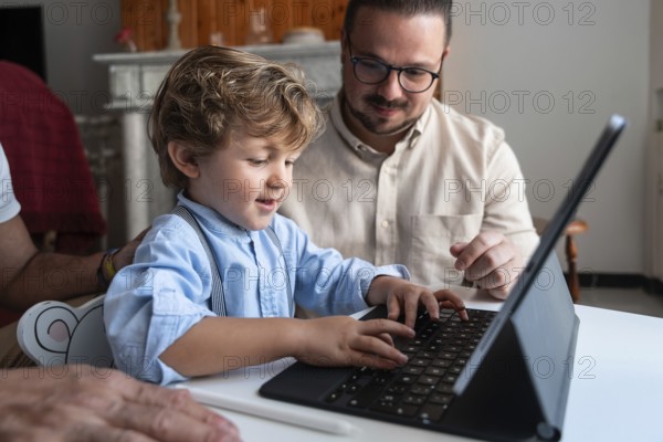 A young child types on a tablet keyboard, guided by an adult. They are sitting together at a table in a cozy home environment, fostering learning and technology skills