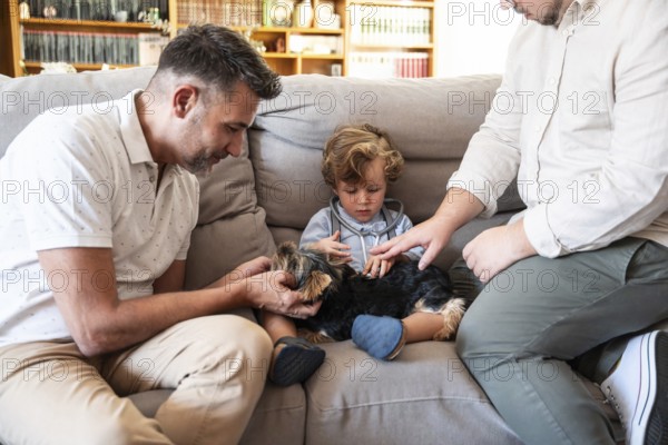 A loving gay couple interacts with their young son and small dog on a cozy couch in their living room
