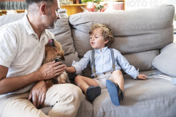 A joyful father and son duo share a warm moment with their small dog on a cozy sofa. The toddler, in suspenders, reaches out to pet the playful canine