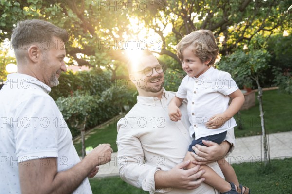 A joyful gay couple shares a tender moment with their son in a sunlit garden, surrounded by lush greenery. The image radiates warmth, love, and family bonding