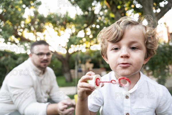 A young boy in a white shirt joyfully blows bubbles in a vibrant garden. His parent, watches in the background, capturing a moment of playful innocence