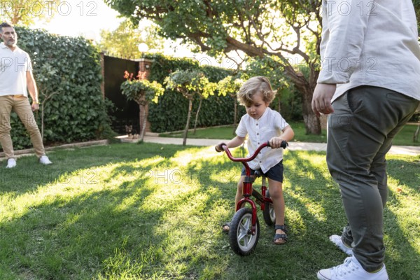 A joyful gay couple encourages their young son as he learns to ride a bicycle in a sunny garden. Lush greenery surrounds this heartwarming family moment