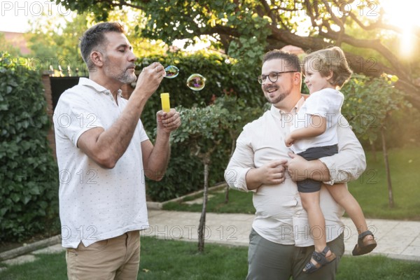 A heartwarming scene shows a gay couple cherishing playful moments with their son outdoors. One parent blows bubbles while the other holds their smiling son