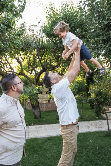 A loving gay couple enjoys quality time with their cheerful son in a lush garden setting. One parent lifts the child playfully as the other watches with joy