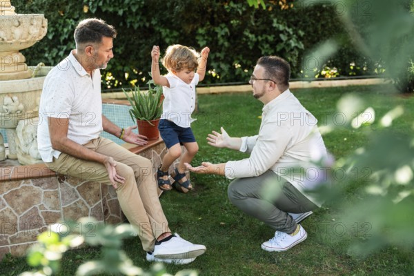 Joyful scene of a gay couple with their son in a lush garden. One father encourages their jumping child, while the other sits nearby, all sharing smiles in the sunlight