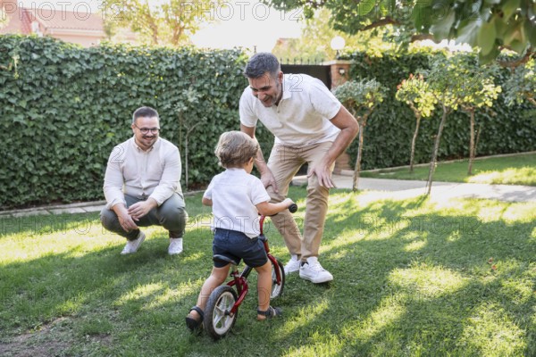 A joyful gay couple plays with their son, teaching him to ride a small bicycle in their garden. The child is learning and having fun under their warm guidance