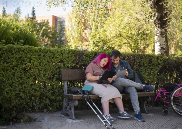 A couple studies together on a bench in a college garden The woman, with pink hair, uses a wheelchair and crutches due to spina bifida, embracing diversity and inclusion