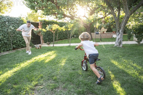 A cheerful scene of a father playing with his young son learning to ride a bicycle in a sunlit garden. The atmosphere is filled with warmth and laughter