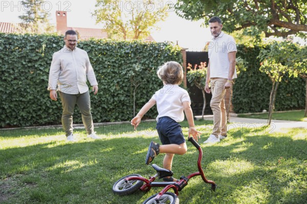 A loving gay couple shares a joyful moment with their young son playing with bicycle in the garden. The sunlight creates a warm atmosphere, highlighting their family bond