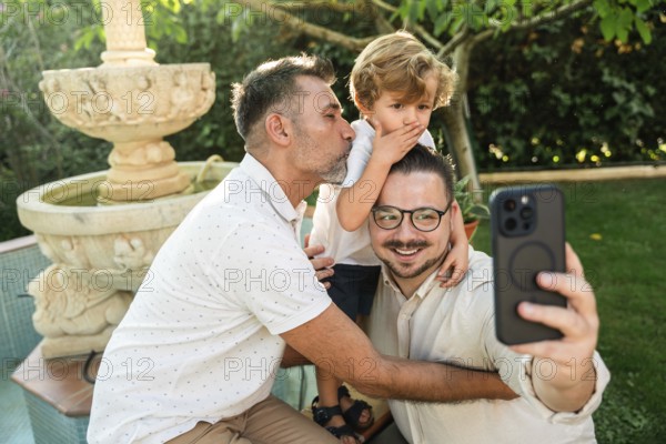 A joyful gay couple takes a selfie with their young son in a lush garden. One dad kisses the son lovingly. The scene radiates warmth and togetherness
