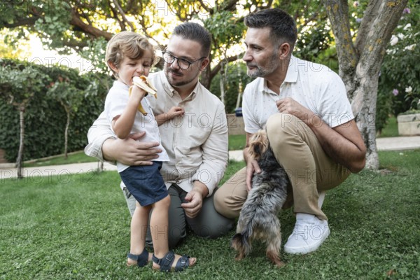 A joyful family moment in a lush garden with a gay couple watching their son eating a banana, and a small dog joining the fun. Love and togetherness in nature