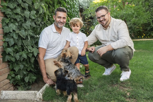 A joyful image of a gay couple with their son and two small dogs outdoors. They are surrounded by greenery, smiling, and enjoying a lovely day in the garden