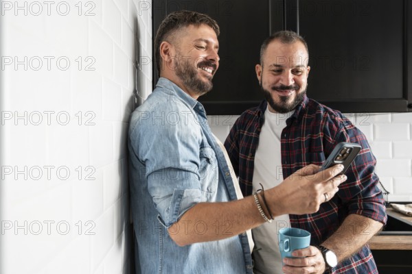 A gay couple enjoying a joyful moment in their kitchen while looking at a smartphone. One holds a blue mug, and both are casually dressed, sharing smiles and warmth