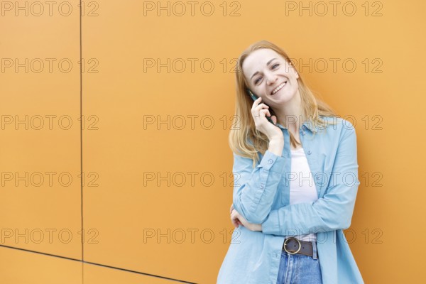 A cheerful woman stands against a bright orange wall in Warsaw, chatting on her phone She's dressed in a casual blue shirt and denim, enjoying a lighthearted moment