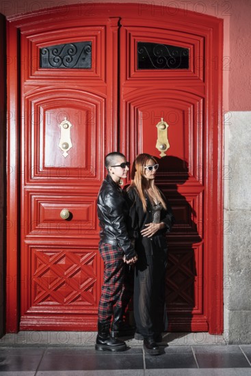 A fashionable lesbian couple stands lovingly against a bright red door, embracing each other's presence in stylish black and red outfits, symbolizing unity and diversity