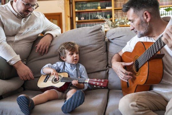 A loving gay couple sits on a sofa, teaching their young son to play the guitar. The warm, family-oriented moment captures joy and togetherness at home