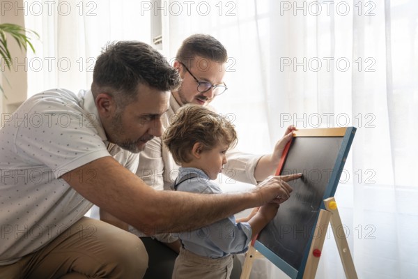 A loving gay couple engages with their young son, teaching him using a chalkboard. The warm, supportive atmosphere captures the essence of family bonding at home