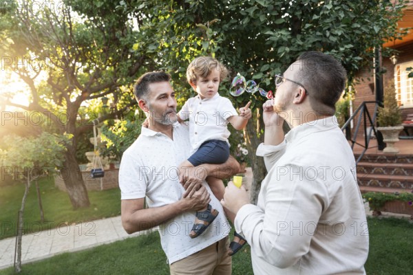 A joyful gay couple spends a sunny afternoon outdoors with their young son, engaging in playful activities, surrounded by lush greenery in their backyard