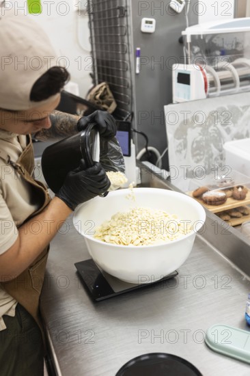 A baker at an Italian bakery carefully pours white chocolate chips into a bowl to weigh it The setting is a professional kitchen with utensils and baked goods in the background