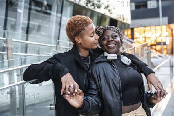 A joyful LGBT multiethnic couple shares a tender moment outdoors. They hold hands and smile warmly, dressed stylishly in casual black attire, with bright city lights in the background